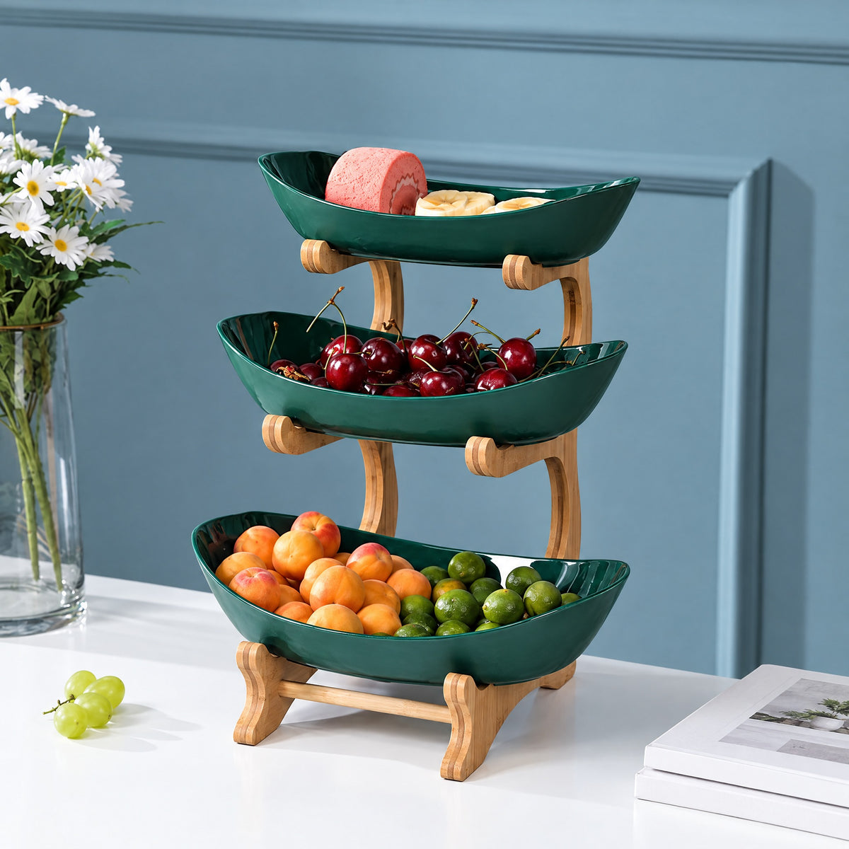 Three-tier fruit stand with deep green ceramic bowls on a natural bamboo frame, holding fruit on a kitchen counter